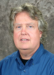Hudmon, Andy 2017-08-15 www Headshot of Andy Hudmon wearing a blue dress shirt in front of a gray photo backdrop.
