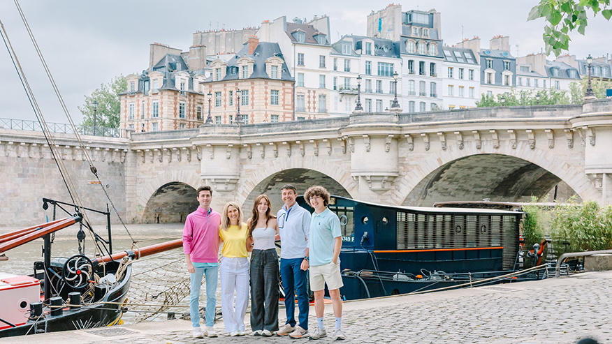 Greg and Amy Baker stand with their two sons and one daughter in front of a gondola and bridge in a European city.