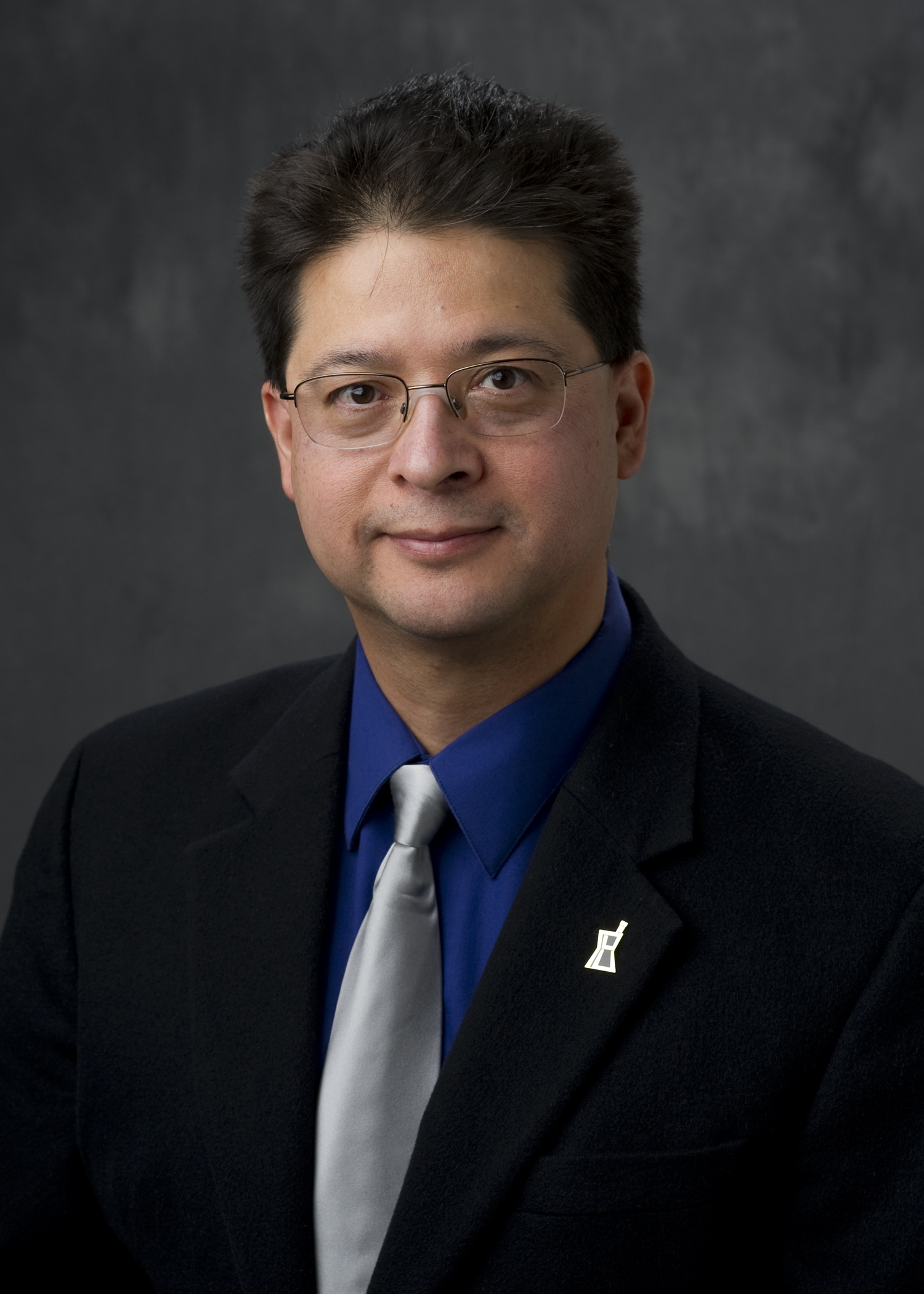 Headshot of Rodolfo Pinal wearing a blue shirt, black blazer and silver tie in front of a dark gray photo backdrop.
