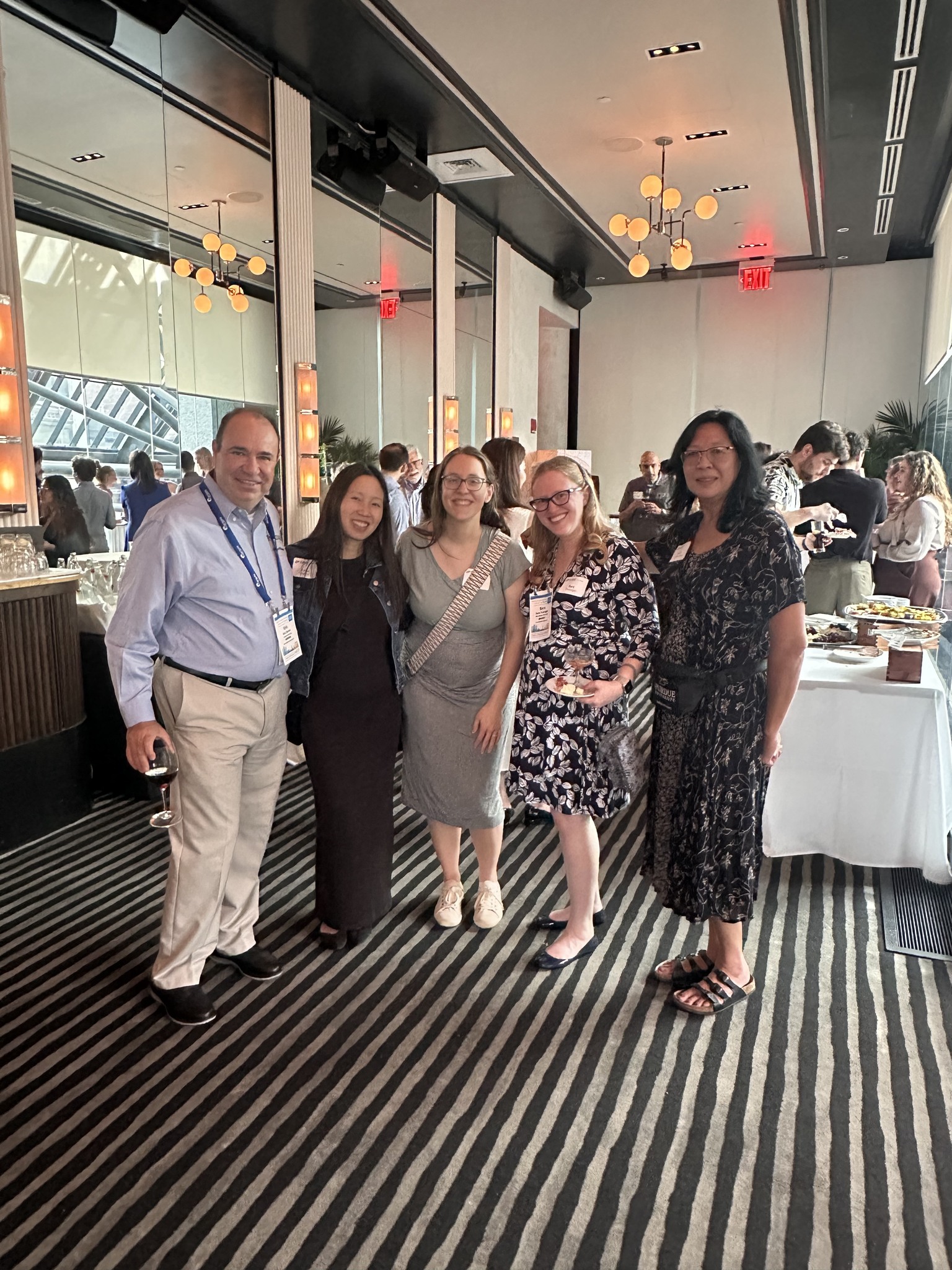 A group of 5 people in dress clothes stand together and smile for the camera at a networking reception.