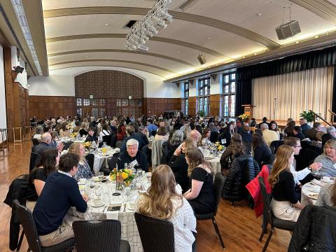 Wide shot of a ballroom with people sitting at round tables with white table cloths in a banquet setting.
