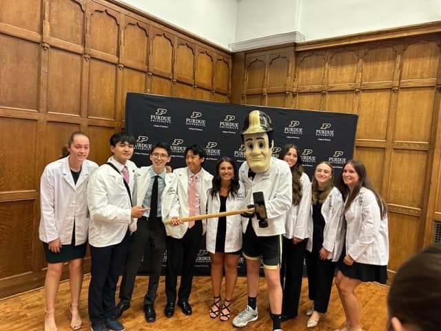 A group of pharmacy students stand in front of a wooden wall with a black Purdue backdrop. They are surrounding Purdue Pete.