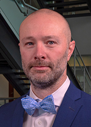 Headshot of Noll Campbell wearing a navy suit jacket, light button-up shirt, and medium blue bowtie.