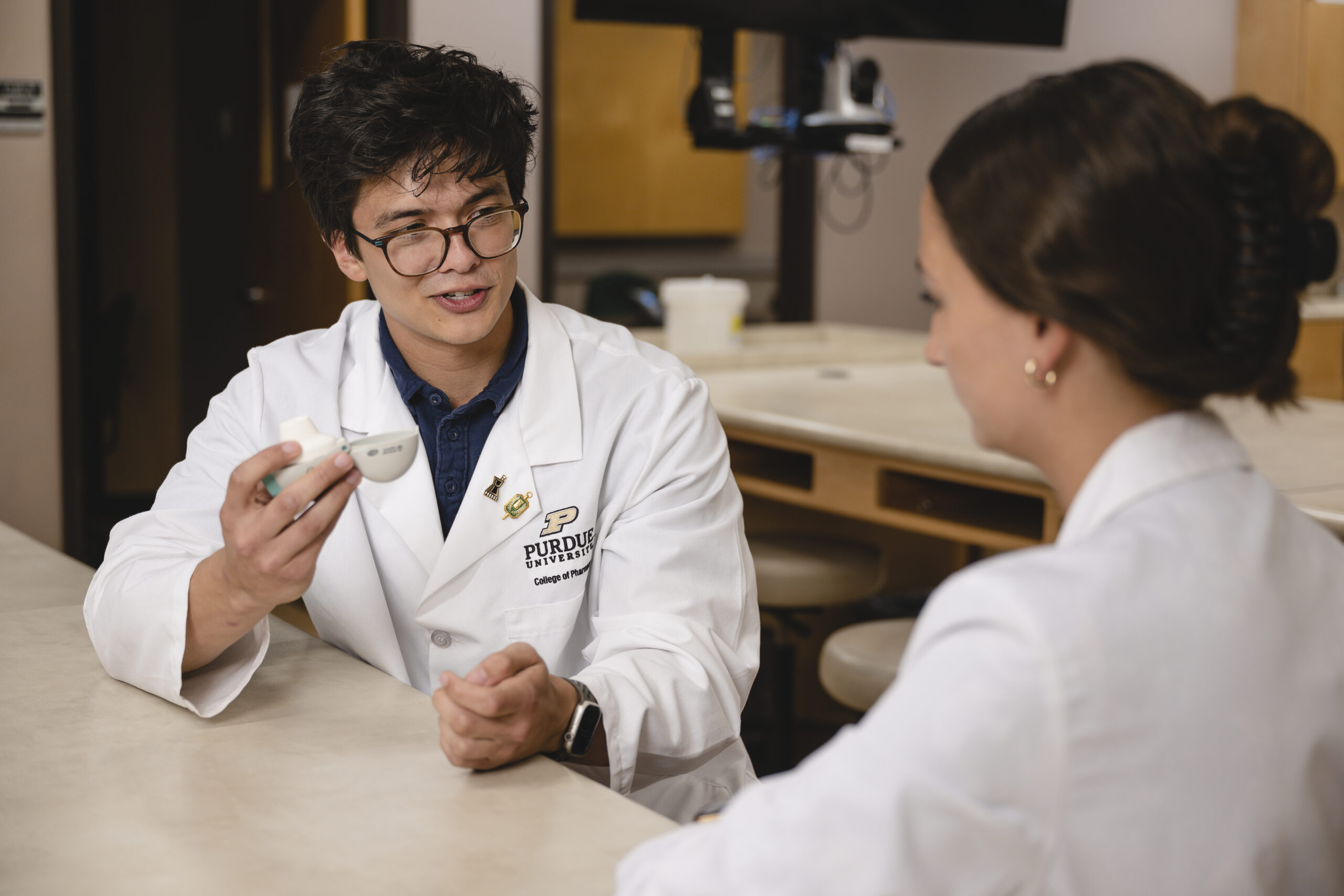 CJ9_8278 A male with glasses wearing a white coat with Purdue logo and pins holds a medication and talks with a female in a white coat facing away from the camera.