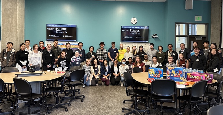 A large group of graduate students stand together in a study area with TVs behind them and study tables in front of them.
