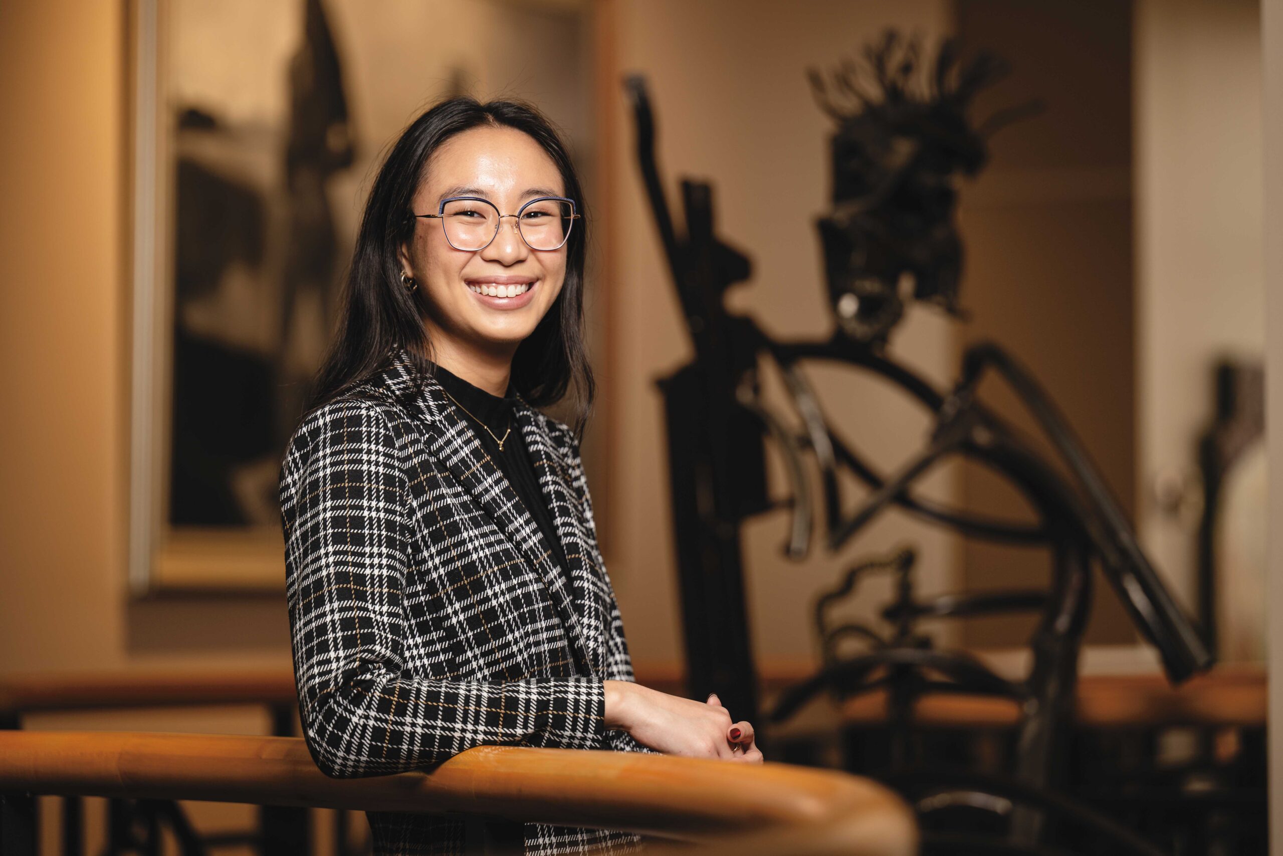 Headshot of Claire Reyes smiling at the camera. She is wearing a black, tan, and white plaid blazer and is leaning on a wooden railing. The backdrop is a blurred background inside Purdue's Black Cultural Center.