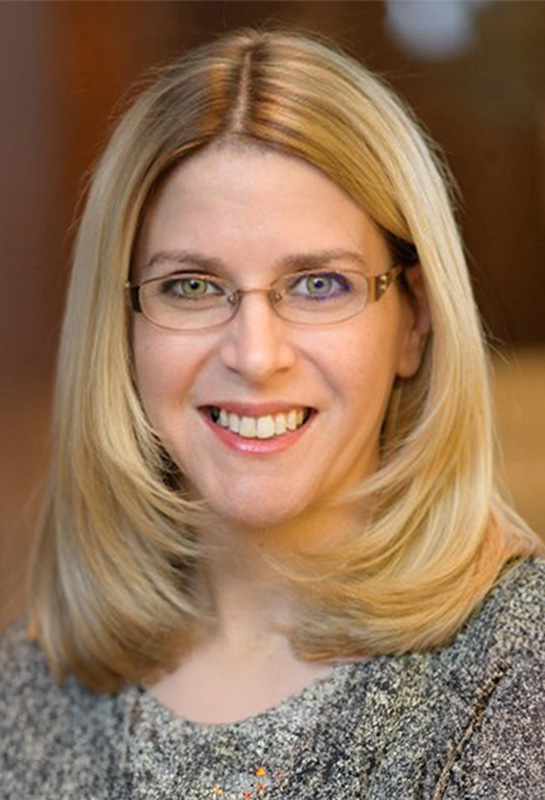 Katie-Skeel-WEB Headshot of Katie Skeel wearing a gray sweater and glasses with a blurred background.