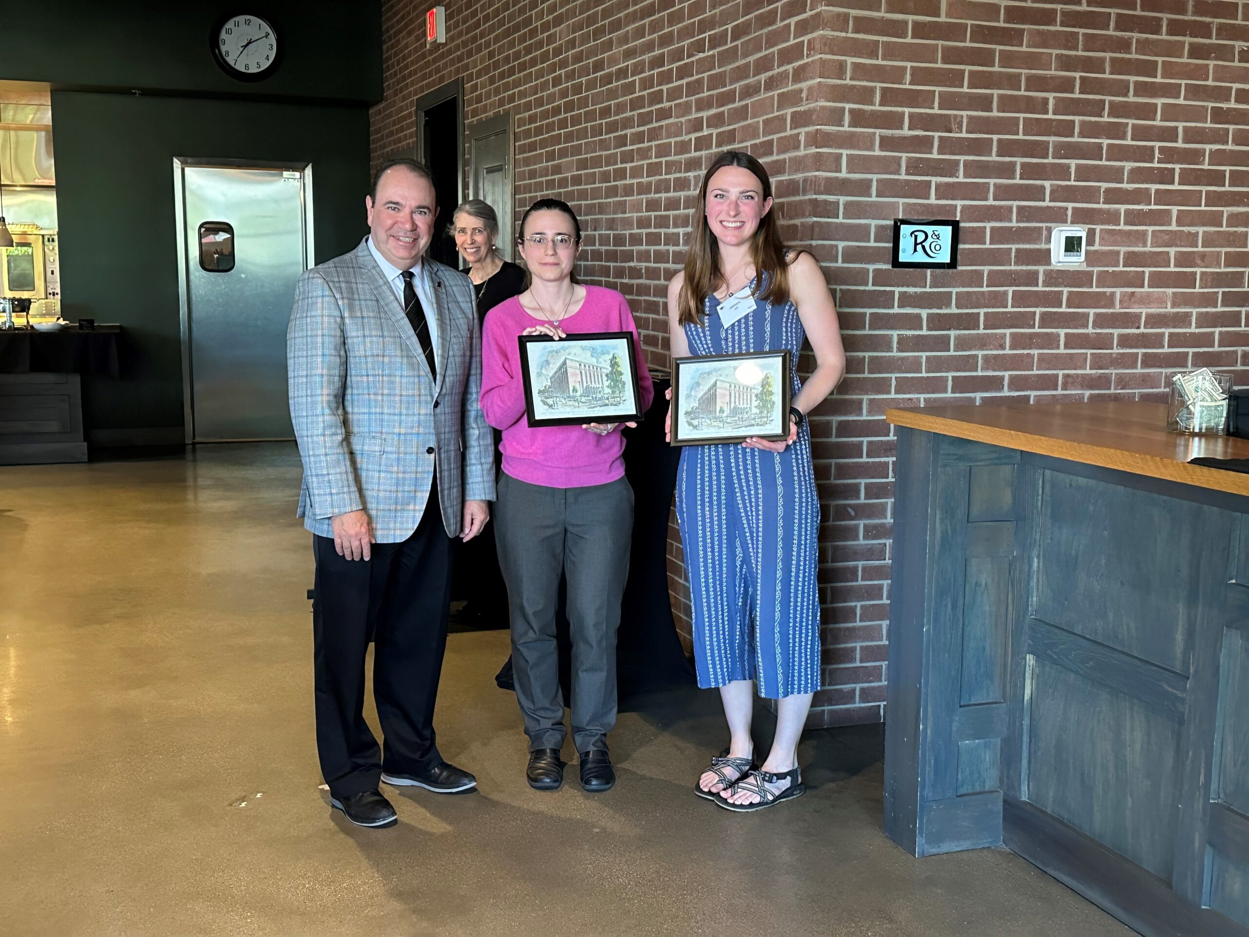 Three people stand indoors against a brick wall, smiling for a photo. Two individuals hold framed artwork, while a third stands beside them. A counter and restaurant interior are visible in the background.