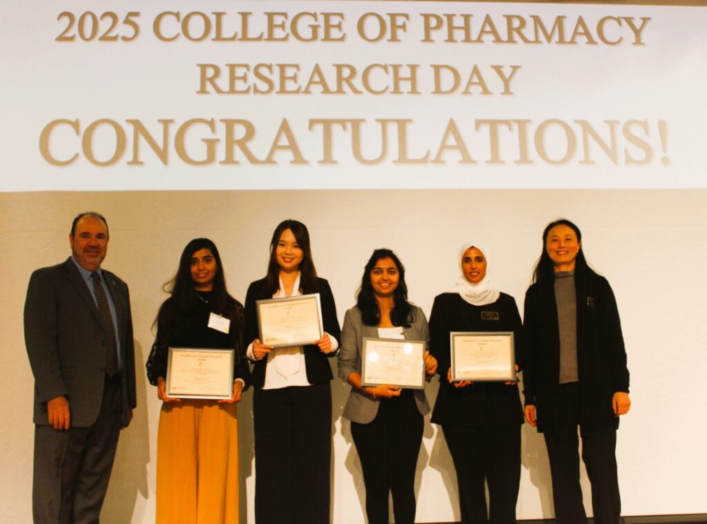 Four students stand between two faculty members in front of a screen. The students are holding awards certificates.