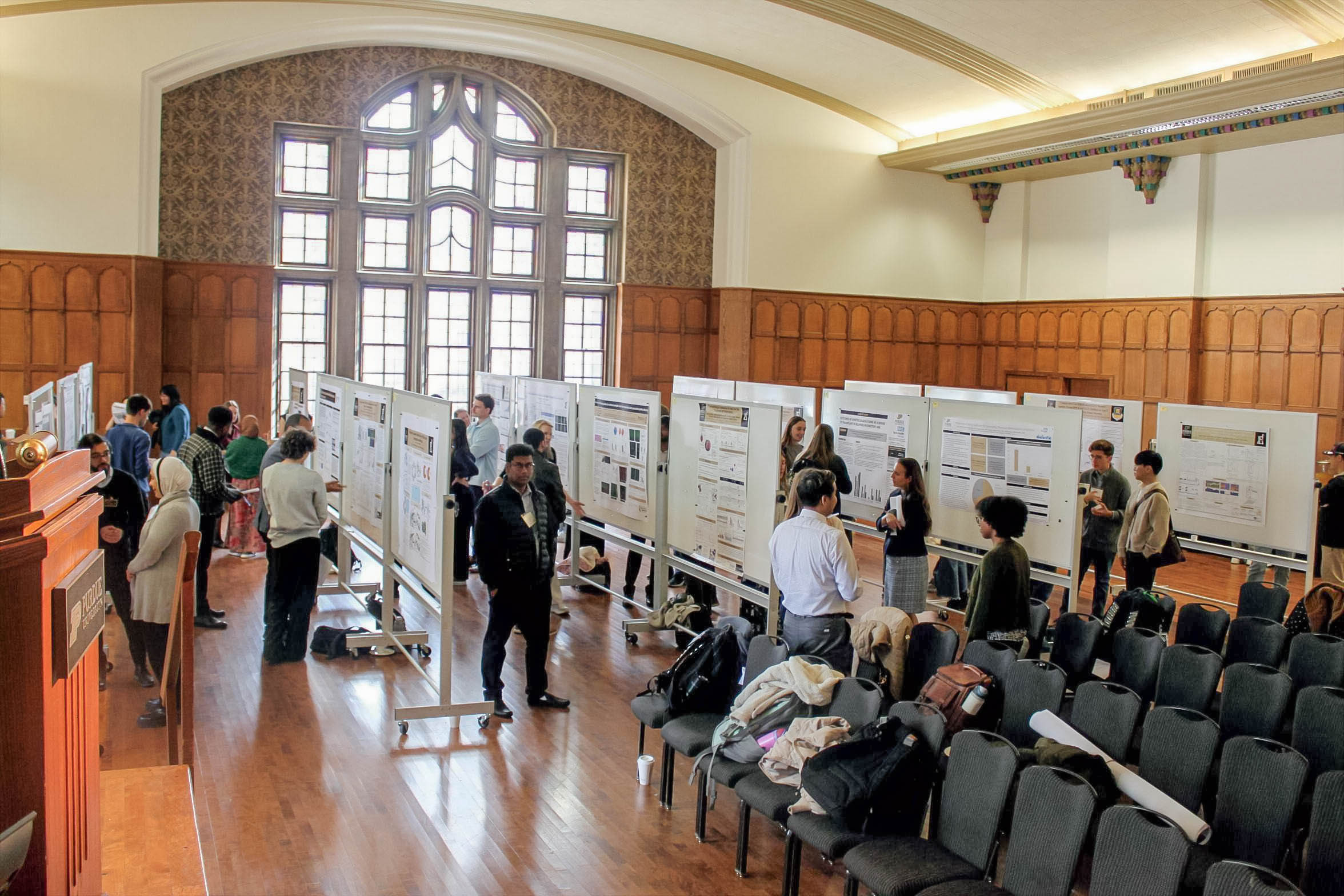 Wide-angled photo of research posters and students looking at them. The setting is the Purdue Memorial Union, with wood floors, wood and white walls, and a floor-to-ceiling ornate window.