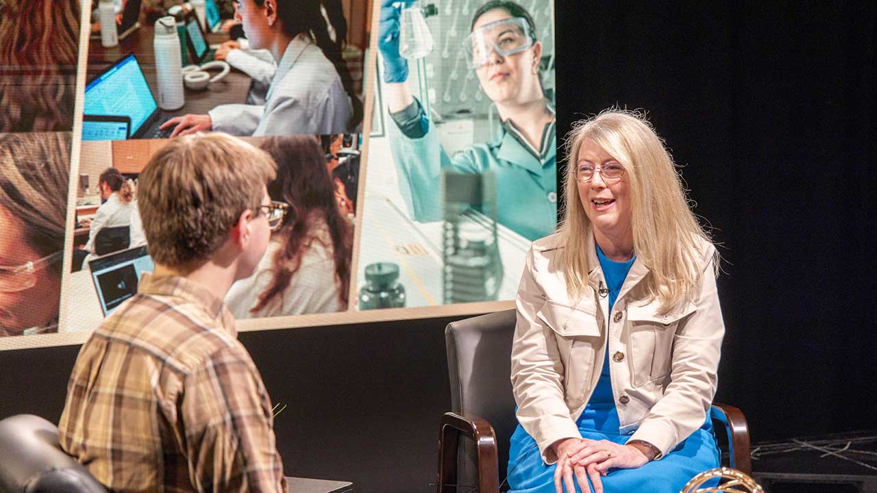 Melanie Dodd, a blonde woman in glasses with blue dress and white jacket, sits in a TV studio with black and screen backdrop. She smiles at host Alex McQuade, who is wearing a plaid dress shirt and facing away from teh camera. Part of the backdrop is a screen with decorative pharmacy images.