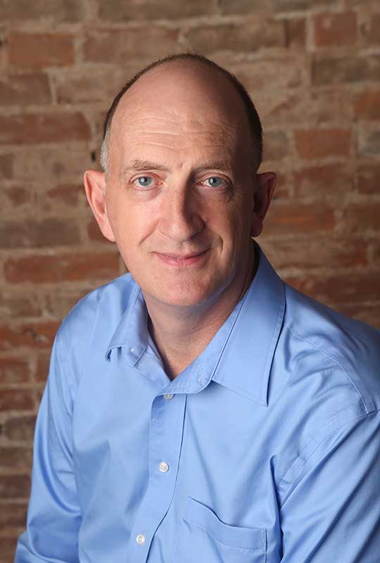 Headshot of Al Thompson looking at the camera. He is wearing a light blue professional shirt. The background is a brick wall.