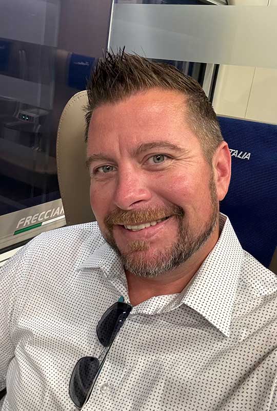Headshot of Brian Schumer on what appears to be an airplane. He is wearing a white collared shirt and smiling at the camera.