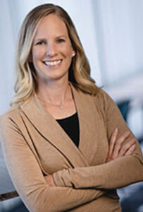Brooke Beier, a woman with shoulder-length blonde hair, smiles at the camera with her arms crossed, wearing a tan cardigan over a dark top, indoors with a softly blurred background.