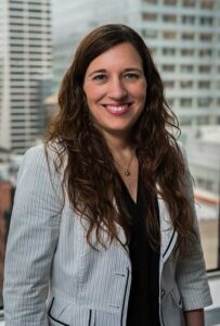 Headshot of Brooke Huang, looking at the camera and wearing a black blouse with white pinstriped blazer. The backdrop is a blurred cityscape of tall buildings.