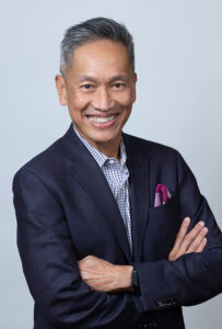 Headshot of Chito Hernandez smiling at the camera. He wears a navy blue suit with a professional shirt and maroon pocket square. The background is a gray photo backdrop.
