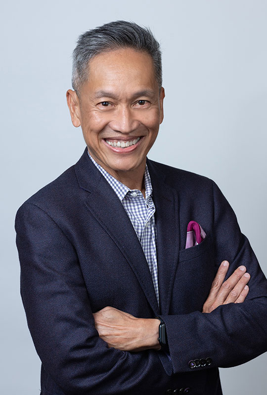 Headshot of Chito Hernandez smiling at the camera. He wears a navy blue suit with a professional shirt and maroon pocket square. The background is a gray photo backdrop.