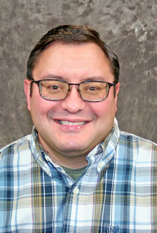 Headshot of Nicholas Duley, wearing glasses and a plaid button-down shirt, smiling against a neutral background.