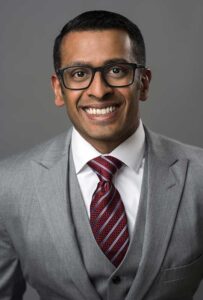 Headshot of Girish Kaimal in front of a medium gray photo backdrop. He wears a medium gray suit with white shirt and maroon pinstriped tie.