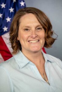 Headshot of Karen Arthur looking at the camera. She is wearing a light blue blouse. The background is a gray photo backdrop and a blurred American flag.