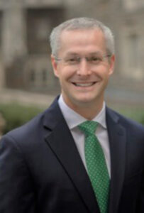 Headshot of Matt Kelm, looking at the camera and smiling. He wears glasses, a dark grey suit, white suit shirt and green tie. The background is a blurred landscape.