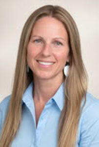 Headshot of Nicole Wallace looking at the camera. She is wearing a light blue professional blouse and the background is an off-white photo backdrop.