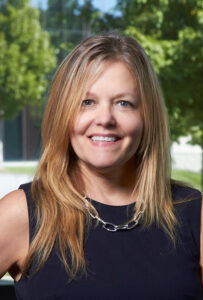 Headshot of Terri Warholak smiling at the camera. She wears a black blouse and the background is a blurred landscape with greenery.