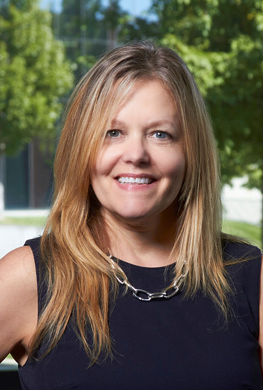 Headshot of Terri Warholak smiling at the camera. She wears a black blouse and the background is a blurred landscape with greenery.