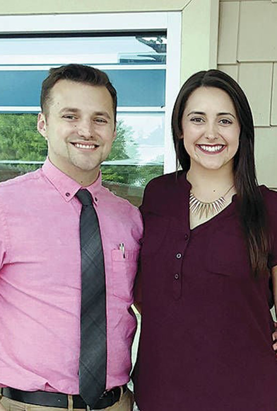 Two adults standing side by side and smiling at the camera outside a building. One wears a pink button-down shirt with a dark tie, and the other wears a dark short-sleeve top and a statement necklace.