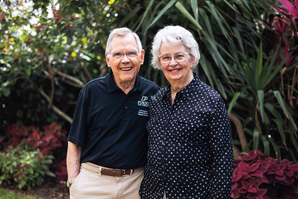 Rick and Anne Borch stand close together outdoors in a lush garden, smiling at the camera. Rick wears glasses and a black Purdue University polo shirt, and Anne wears glasses and a black blouse with a small white pattern, with greenery and colorful plants in the background.