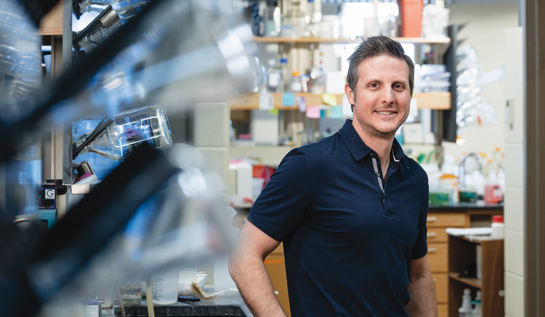 A researcher stands in a laboratory, smiling toward the camera. He is wearing a navy short-sleeve polo shirt and is positioned beside a lab bench, with shelves, glassware, and scientific equipment visible in the softly blurred background.