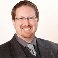 Headshot of Jonathon Parker wearing glasses and smiling at the camera. He wears a black suit, gray dress shirt, and light tie.