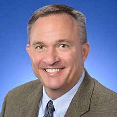 Headshot of James Wesley in a gray blazer and tie with light blue dress shirt. He smiles in front of a blue photo backdrop.