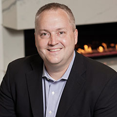 Headshot of Joseph Trebley smiling at the camera with a fireplace in the background. He wears a lavender dress shirt with black suit jacket.