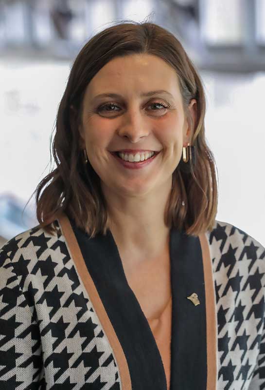 Headshot of Jennifer Stewart-Burton smiling for the camera in front of a bright blurred background. She wears a tan shirt with black and white houndstooth sweater with tan pinstripe.
