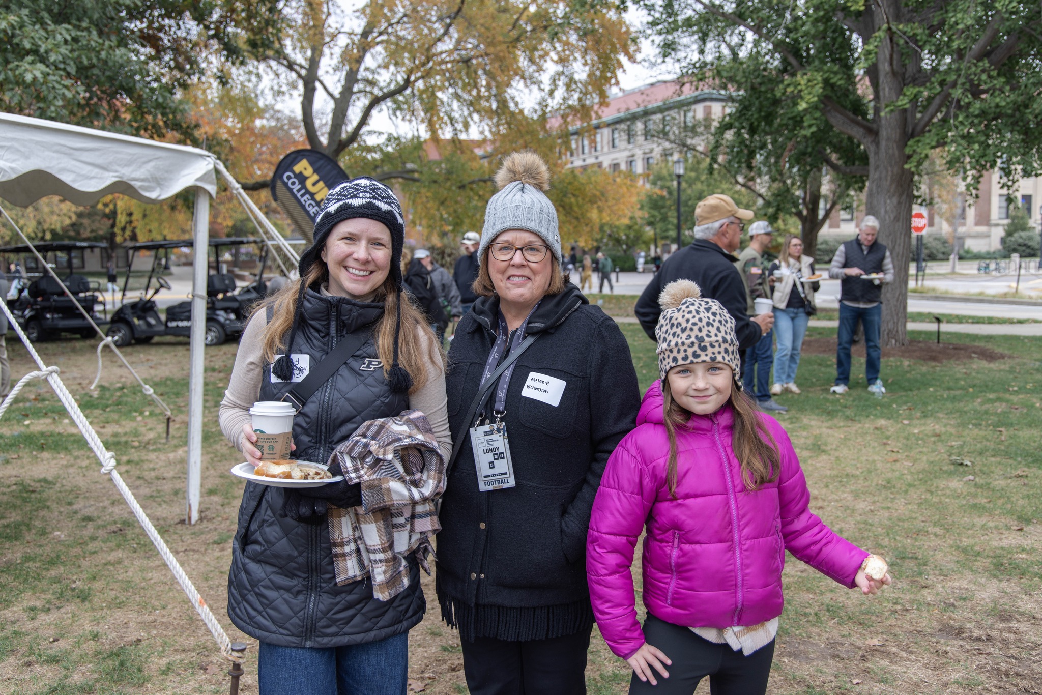 Two women and a girl stand together smiling at the camera. The background is memorial mall and other attendees of a tailgate.
