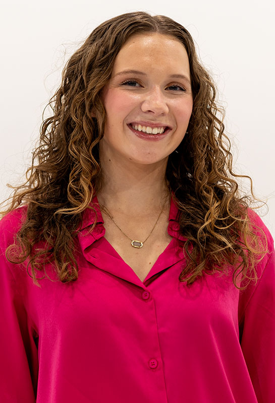 Headshot of Ashley Jensen smiling at the camera and wearing a pink blouse in front of a white photo backdrop.