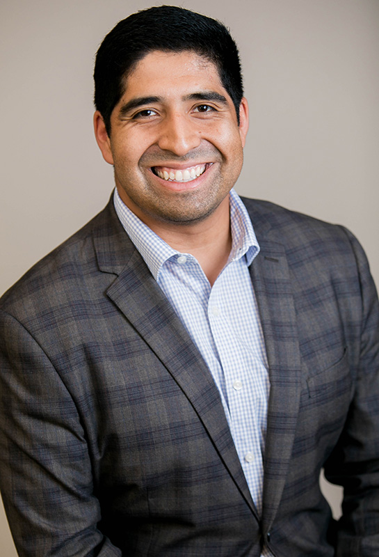 Headshot of Devin Rittenhouse wearing a suit and smiling for the camera in front of a blank photo backdrop.