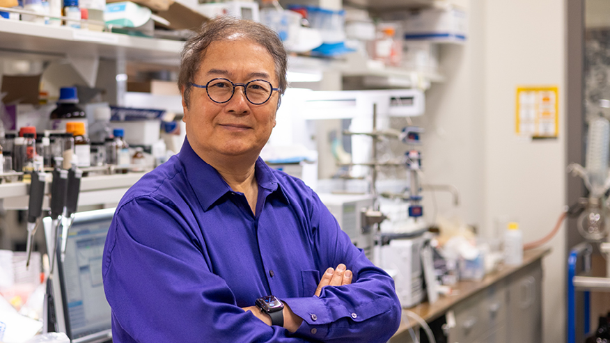 Headshot of Zhong-Yin Zhang in a dark blue shirt with arms crossed looking at the camera. The background is a laboratory bench.