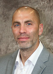 Headshot of Ryan Altman looking at the camera. He wears a white dress shirt and gray suit jacket in front of a textured gray photo backdrop.